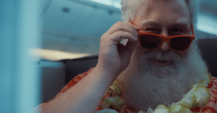 Le Père Noël dans le nouveau film d'United Airlines, l'amour en vue d'un avion.  Il porte une chemise hawaïenne, des lunettes de soleil et un collier.  Il est assis en classe affaires et fait un clin d'œil.